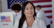 © vesperstock - CU Mature woman in white lab coat with 'I Voted' sticker. Other scientists/doctors of various demographics, in white lab coats, vote in background