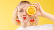 © Вячеслав Косько - Stock photo of positive blonde young woman in white t-shirt with halved orange holding it in front of her eye and pouting lips at camera. Isolate on yellow background. Summer concept.