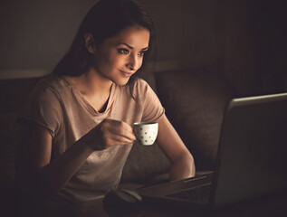 Smiling thinking beautiful woman looking on computer monitor drinking coffee on dark shadow home background in the evening.