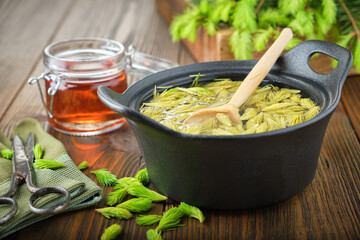 Wall Mural - Cooked spruce tips in a black pan, jar of  honey or syrup from fir buds and needles, twigs of fir tree on wooden table. Making Spruce tips jam at home.