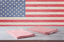 Empty Wooden Table With Striped Tablecloth Over Brick Wall With American Flag. 4th Of July USA Independence Day Mock Up For Design.