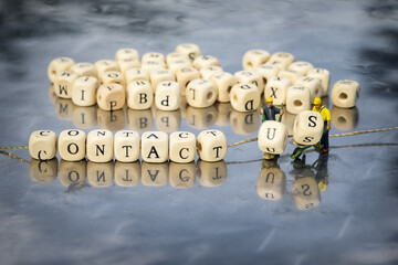 Miniature model of people and wooden cubes with contact us inscription strung on a thread on reflective table. Construction workers with wheelbarrows add cubes u and s.