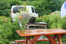 
A Bouquet Of Wildflowers Stands On A Table Wet From Rain. A White Minibus Stands Near Green Bushes In The Background