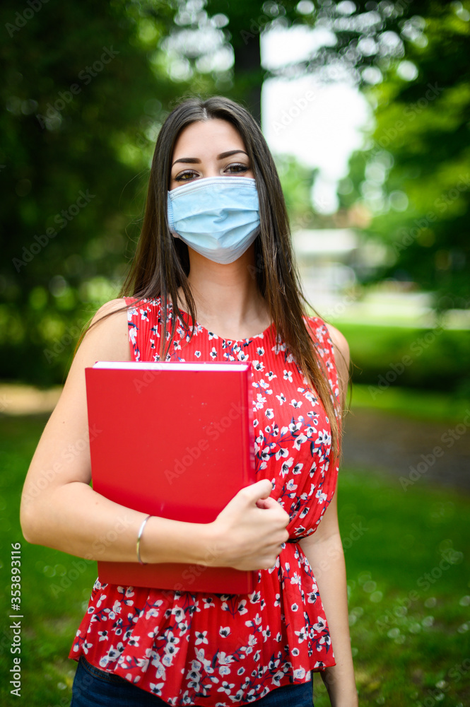 Beautiful female student holding a book outdoor and wearing a mask, coronavirus concept - obrazy, fototapety, plakaty 
