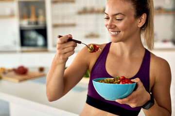 Happy athletic woman having fruit salad for a snack in the kitchen.