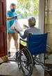 A man wearing a mask working with a church group or other benevolent organization brings some groceries to an elderly woman in a wheelchair.
