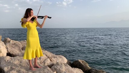 Wall Mural - Young beautiful musician girl in yellow dress is playing electric violin on rocks near Mediterranean sea. Open air solo concert concept. 4k stock video.  