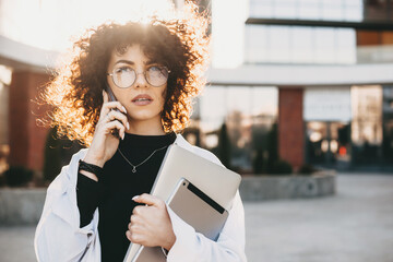 Young entrepreneur with curly hair and eyeglasses talking on phone while posing with a laptop outside