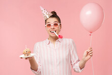 Young Happy Birthday Girl Wearing Colored Glasses And Holiday Hat, Holding Balloon, Cake And Whistle In Mouth, Isolated On Pink Background