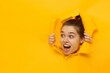 Young excited girl looking through hole in paper at beneficil commercial offer with eyes round with surprise, isolated on yellow background with copy space