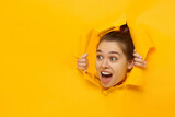 Fototapeta  - Young excited girl looking through hole in paper at beneficil commercial offer with eyes round with surprise, isolated on yellow background with copy space