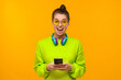 © Damir Khabirov - Young teenage girl in green neon top and eyeglasses, wearing blue wireless headphones around neck, holding smartphone in hands and feeling excited and shocked, isolated on yellow background