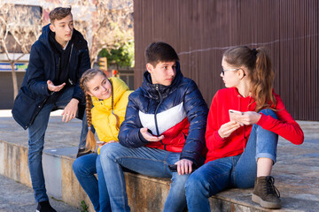  Four teenagers enthusiastically look at the screens of their smartphones on street