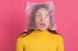 Shocked female wearing yellow shirt posing with plastic bag on her head, looks shocked and astonished, keeping mouth widely opened, stands against rose wall.