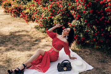 sensual woman with dark hair in luxurious dress and accessories posing in summer garden with red roses