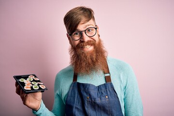 Sticker - Redhead Irish cook man with beard holding maki sushi tray over isolated background with a happy face standing and smiling with a confident smile showing teeth