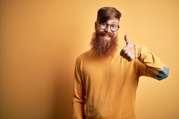 Canvas Print - Handsome Irish redhead man with beard wearing glasses over yellow isolated background doing happy thumbs up gesture with hand. Approving expression looking at the camera showing success.