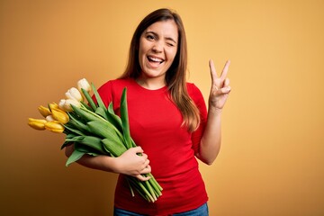 Sticker - Young blonde woman holding romantic bouquet of tulips flowers over yellow background smiling with happy face winking at the camera doing victory sign with fingers. Number two.