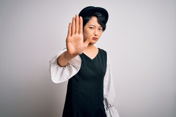 Wall Mural - Young beautiful asian girl wearing casual dress and hat standing over isolated white background doing stop sing with palm of the hand. Warning expression with negative and serious gesture on the face.