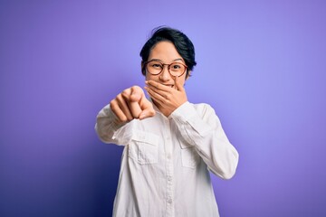 Young beautiful asian girl wearing casual shirt and glasses standing over purple background laughing at you, pointing finger to the camera with hand over mouth, shame expression