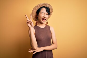 Young beautiful asian girl wearing casual t-shirt and hat over isolated yellow background smiling with happy face winking at the camera doing victory sign. Number two.