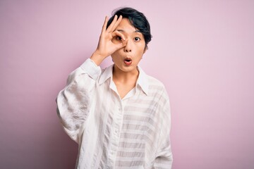 Wall Mural - Young beautiful asian girl wearing casual shirt standing over isolated pink background doing ok gesture shocked with surprised face, eye looking through fingers. Unbelieving expression.