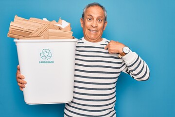 Wall Mural - Senior man recycling holding trash can with cardboard to recycle over blue background with surprise face pointing finger to himself