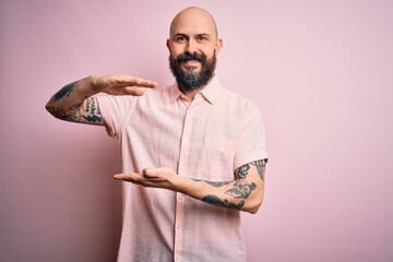 Sticker - Handsome bald man with beard and tattoo wearing casual shirt over isolated pink background gesturing with hands showing big and large size sign, measure symbol. Smiling looking at the camera. Measure