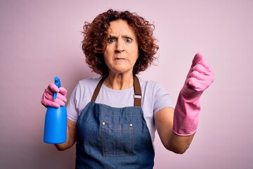 Poster - Middle age curly hair woman cleaning doing housework wearing apron and gloves using spayer annoyed and frustrated shouting with anger, crazy and yelling with raised hand, anger concept