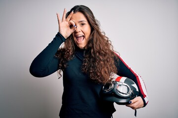 Sticker - Beautiful motorcyclist woman with curly hair holding moto helmet over white background with happy face smiling doing ok sign with hand on eye looking through fingers