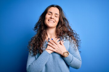 Sticker - Young beautiful woman with curly hair wearing blue casual sweater over isolated background smiling with hands on chest with closed eyes and grateful gesture on face. Health concept.