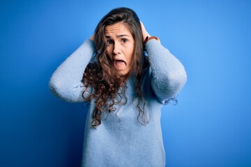 Poster - Young beautiful woman with curly hair wearing blue casual sweater over isolated background Crazy and scared with hands on head, afraid and surprised of shock with open mouth