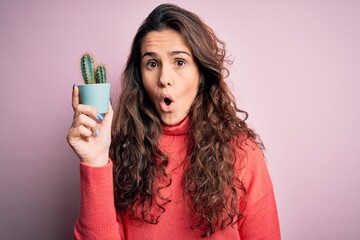 Sticker - Young beautiful woman with curly hair holding small cactus over isolated pink background scared in shock with a surprise face, afraid and excited with fear expression