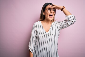 Wall Mural - Young beautiful woman wearing casual striped t-shirt and glasses over pink background very happy and smiling looking far away with hand over head. Searching concept.