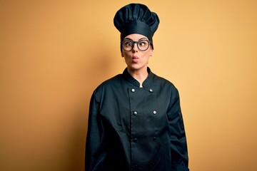 Poster - Young beautiful brunette chef woman wearing cooker uniform and hat over yellow background making fish face with lips, crazy and comical gesture. Funny expression.