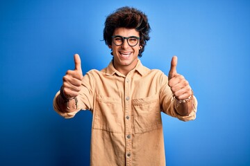 Young handsome man wearing casual shirt standing over isolated blue background success sign doing positive gesture with hand, thumbs up smiling and happy. Cheerful expression and winner gesture.