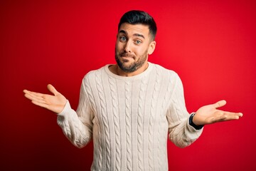 Wall Mural - Young handsome man wearing casual white sweater standing over isolated red background clueless and confused expression with arms and hands raised. Doubt concept.