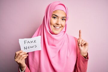 Canvas Print - Young beautiful girl wearing muslim hijab holding paper with fake news message surprised with an idea or question pointing finger with happy face, number one