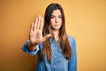 Poster - Young beautiful girl wearing casual denim shirt standing over isolated yellow background doing stop sing with palm of the hand. Warning expression with negative and serious gesture on the face.