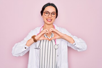 Wall Mural - Beautiful doctor woman with blue eyes wearing coat and stethoscope over pink background smiling in love doing heart symbol shape with hands. Romantic concept.