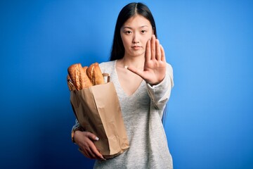 Wall Mural - Young asian woman holding bread grocery paper bag over blue isolated background with open hand doing stop sign with serious and confident expression, defense gesture