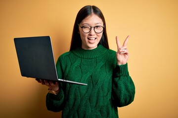 Poster - Young asian business woman wearing glasses and working using computer laptop smiling with happy face winking at the camera doing victory sign with fingers. Number two.