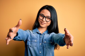 Wall Mural - Young beautiful chinese woman wearing casual denim shirt over isolated yellow background looking at the camera smiling with open arms for hug. Cheerful expression embracing happiness.