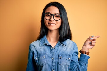 Wall Mural - Young beautiful chinese woman wearing casual denim shirt over isolated yellow background with a big smile on face, pointing with hand finger to the side looking at the camera.