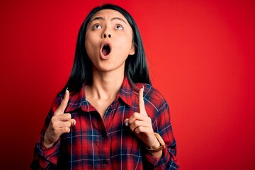 Wall Mural - Young beautiful chinese woman wearing casual shirt over isolated red background amazed and surprised looking up and pointing with fingers and raised arms.