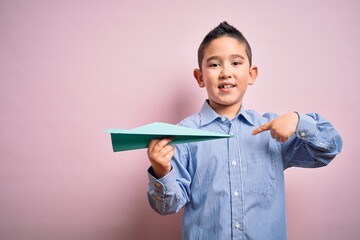 Sticker - Young little boy kid holding paper plane over isolated pink background with surprise face pointing finger to himself