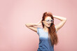 © etonastenka - Portrait of surprised young woman in sunglasses enjoying in front of pink wall. Indoor portrait of redhead lady in sunglasses fooling around in studio