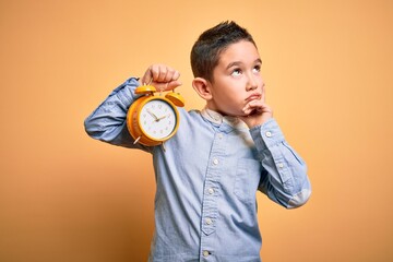 Wall Mural - Young little boy kid holding classic bell alarm clock over isolated yellow background serious face thinking about question, very confused idea