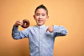 Poster - Young little boy kid eating unhealthy chocolate doughnut over isolated yellow background with surprise face pointing finger to himself