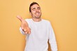 © Krakenimages.com - Young handsome man with blue eyes wearing casual sweater standing over yellow background smiling cheerful offering palm hand giving assistance and acceptance.